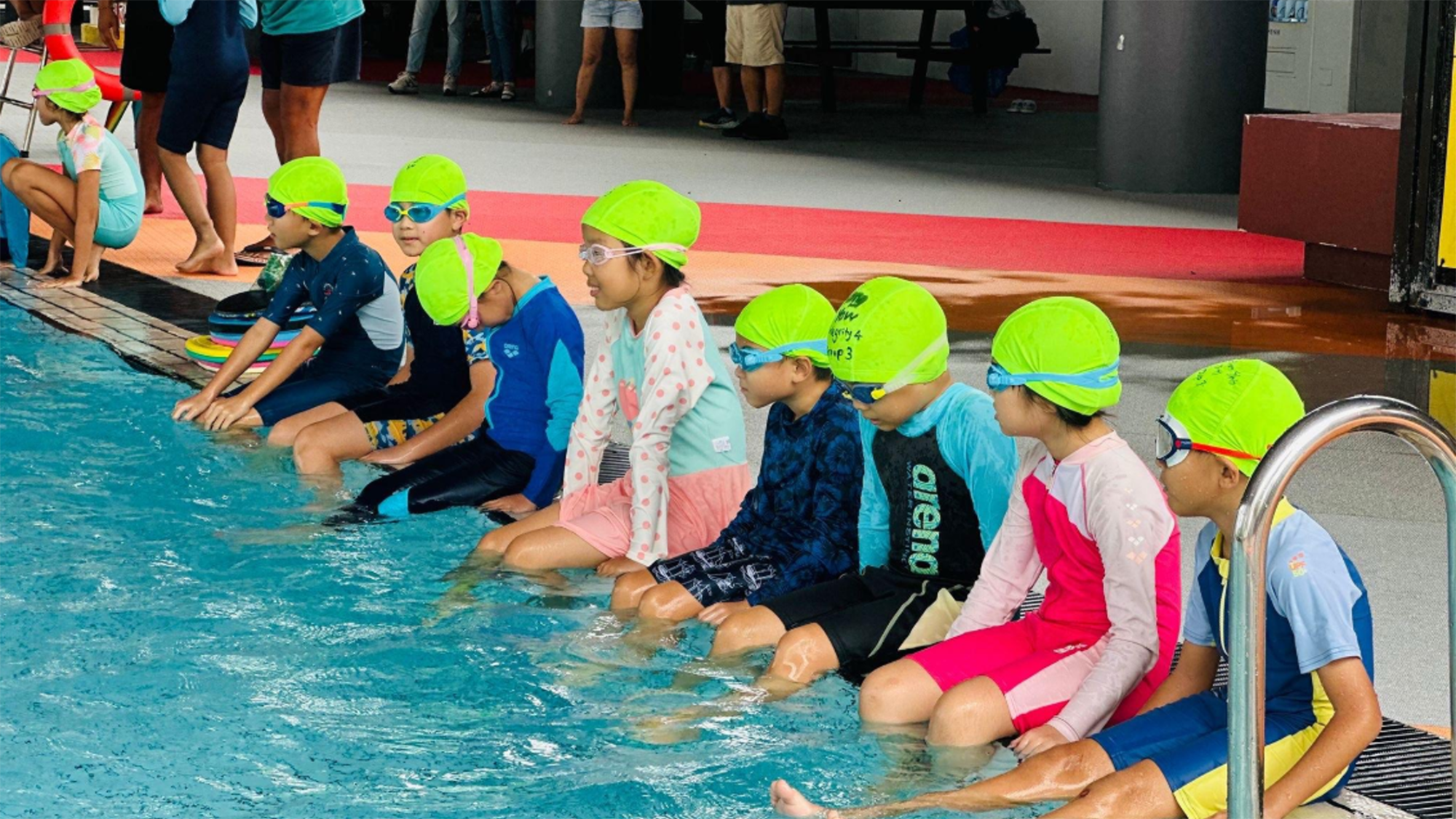 A group of students wearing bright green swim caps and goggles sit at the edge of a swimming pool with their feet in the water as they listen to instructions before their swimming lesson.